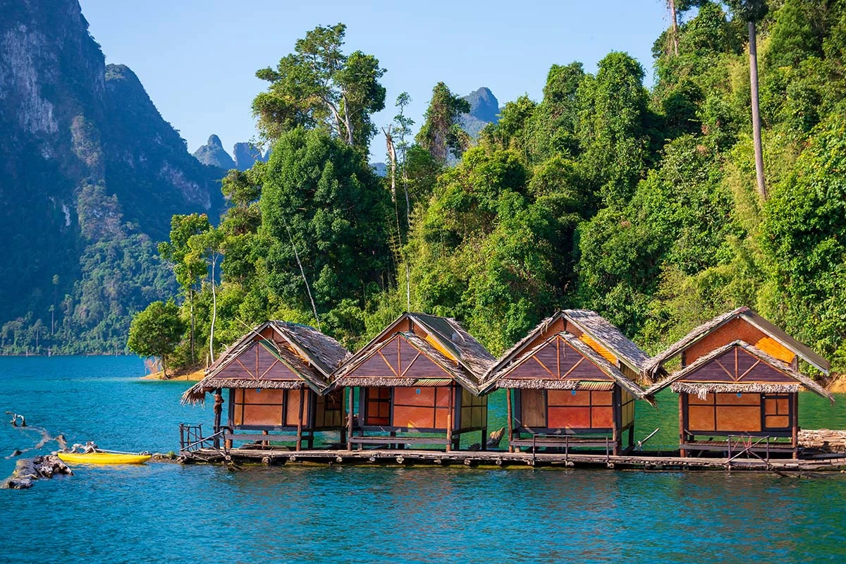 Four wooden floating huts on a blue lake, surrounded by lush green forest and distant mountains.