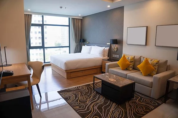 Modern hotel room with a bed, sofa, desk, large window, and decorative pillows and artwork.