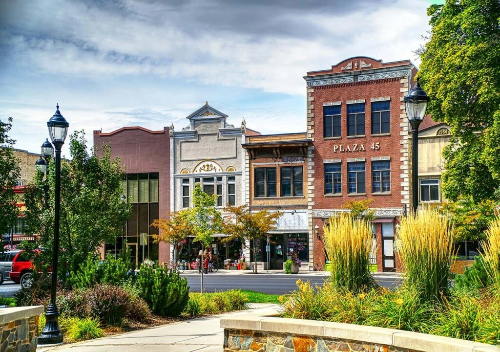 Colorful historic buildings and shops line a small town street, with lush plants and trees in the foreground.
