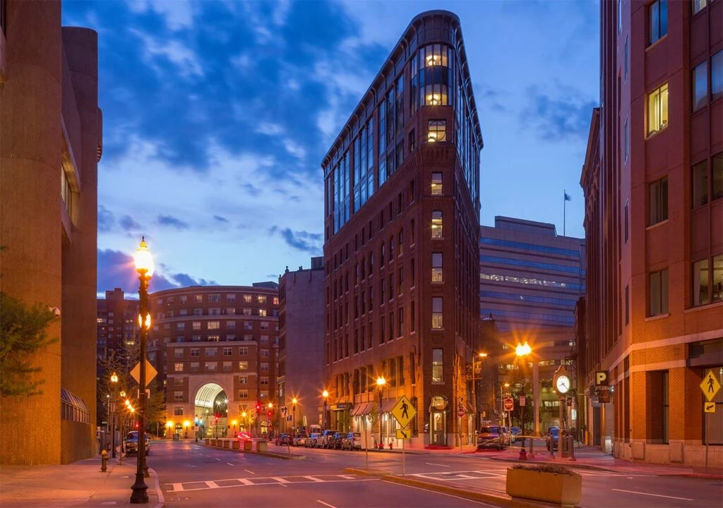 City street at dusk with illuminated buildings, streetlights, and a clear blue sky with scattered clouds.
