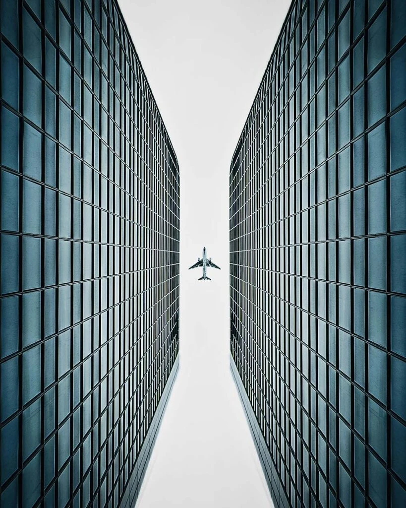 An airplane flies between two tall glass buildings under a clear, pale sky.