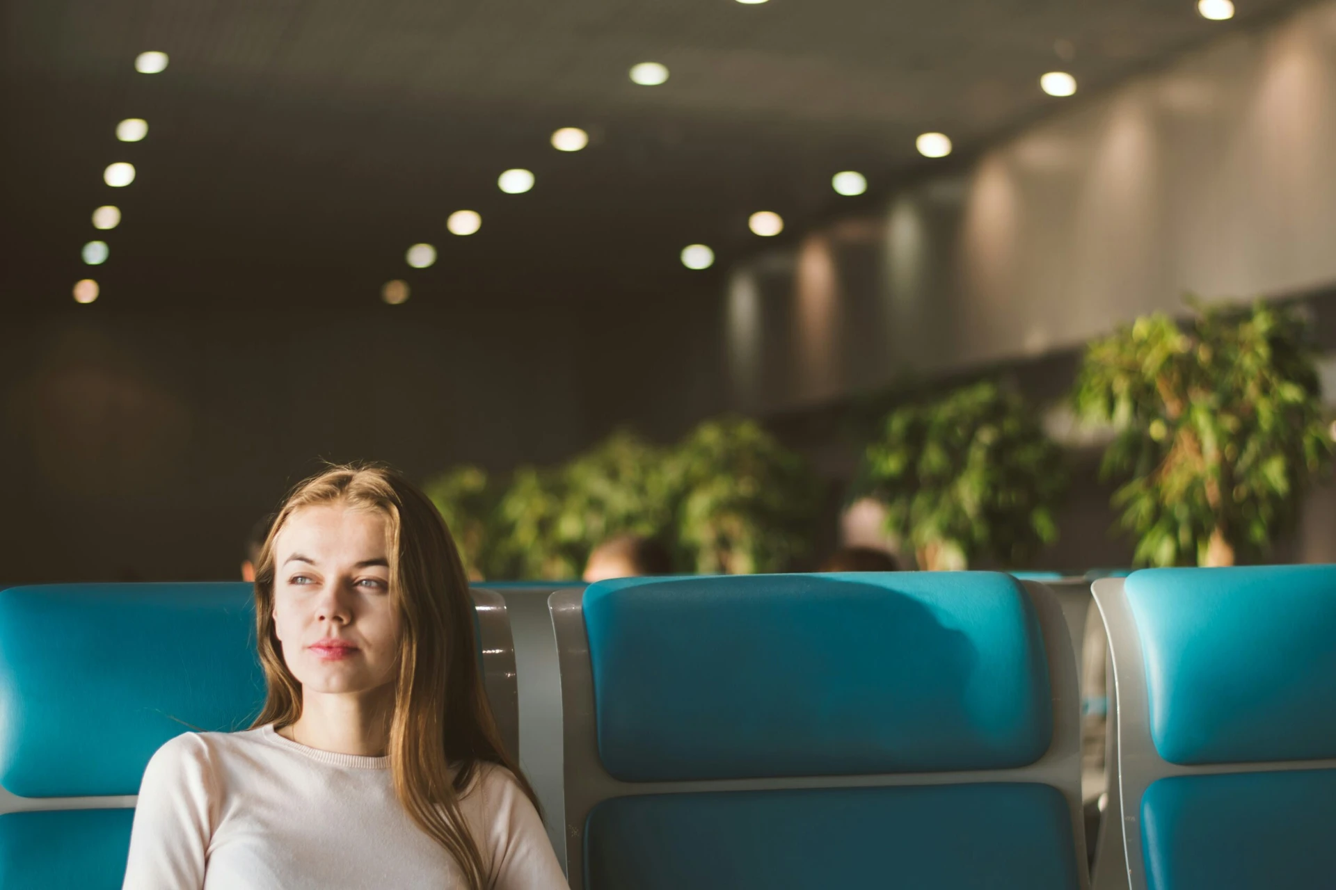 Woman sitting alone on blue seats in a waiting area with green plants and ceiling lights in the background.