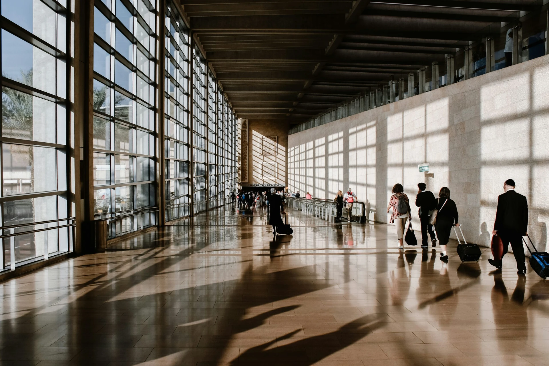 Travelers with luggage walk through a bright, sunlit airport terminal with large windows.