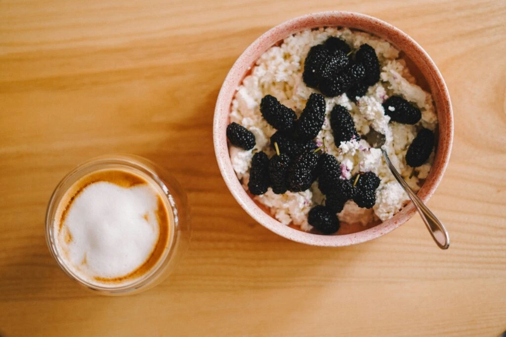 A bowl of cottage cheese with blackberries and a spoon beside a cup of frothy coffee on a wooden table.
