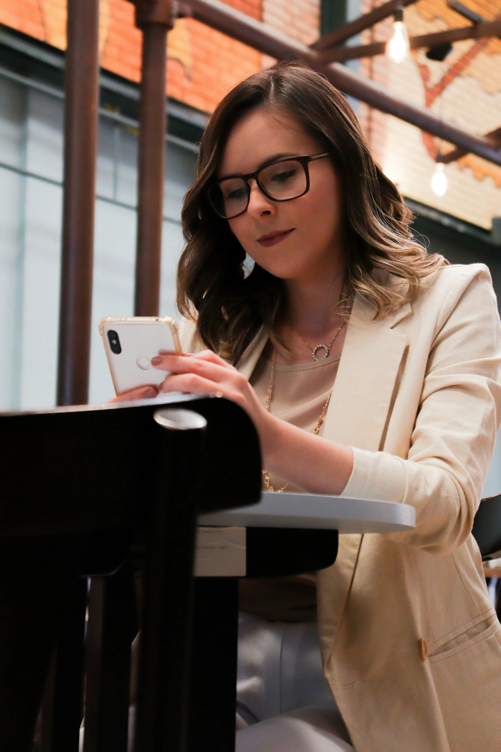 Woman with glasses and beige blazer sits at a table, looking at her phone in a modern indoor setting.