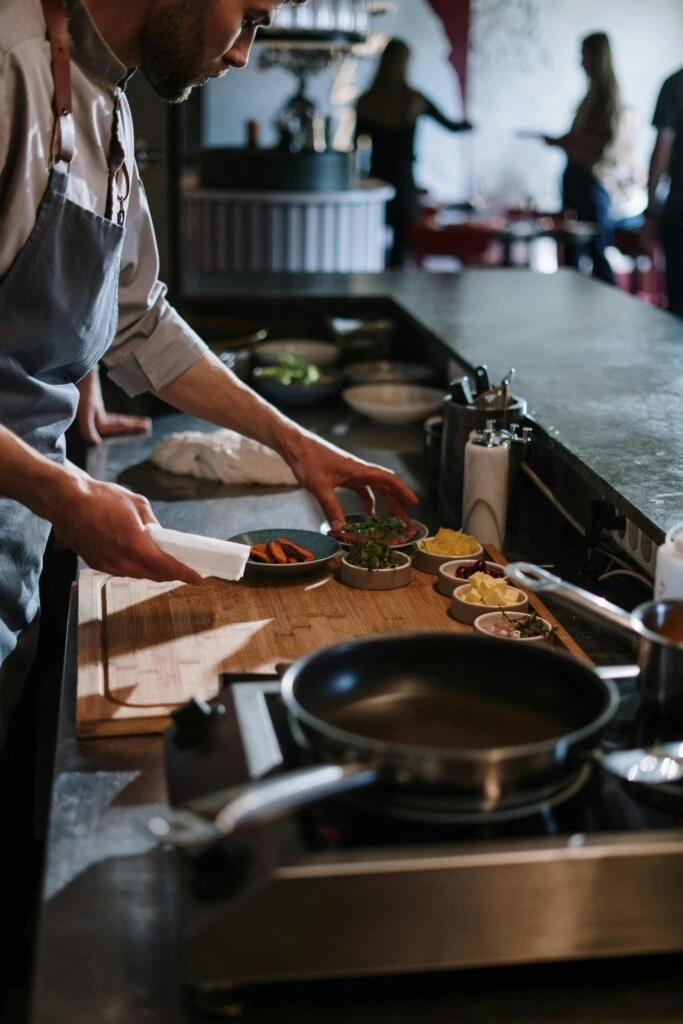Chef preparing ingredients on a wooden board in a restaurant kitchen with pans and bowls nearby.