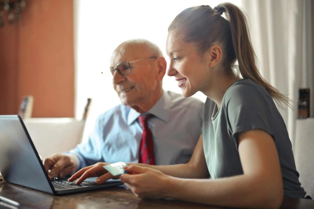 Young woman helps older man use a laptop; both are smiling and looking at the computer screen together.