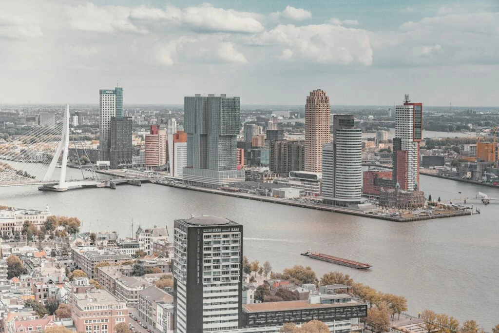 A city skyline with modern high-rises beside a wide river and a white cable-stayed bridge on a cloudy day.