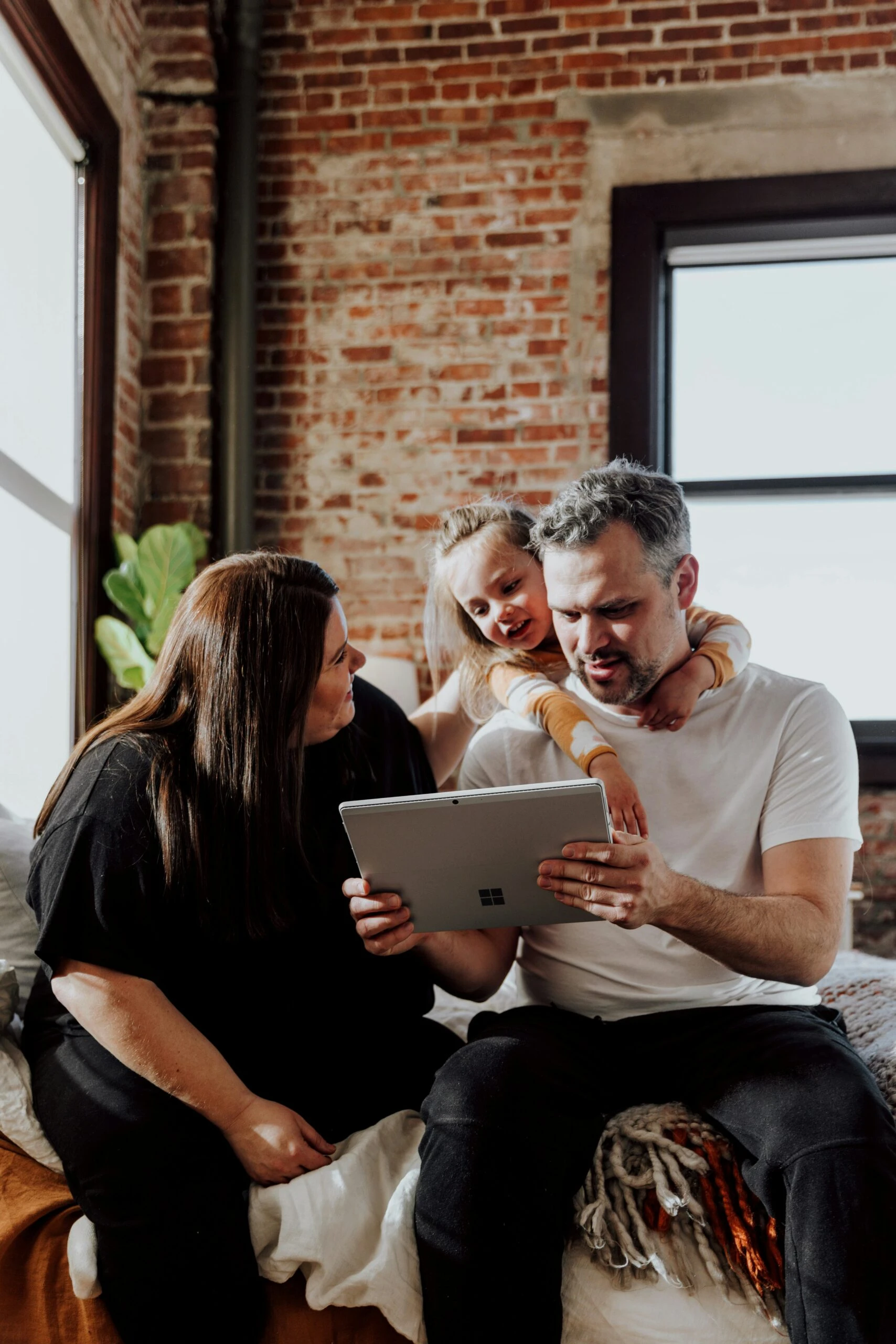 A family of three sits on a bed, smiling and looking at a tablet together in a cozy room.