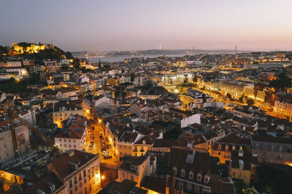 Aerial view of Lisbon at sunset, with city lights, red rooftops, and a river in the distance.