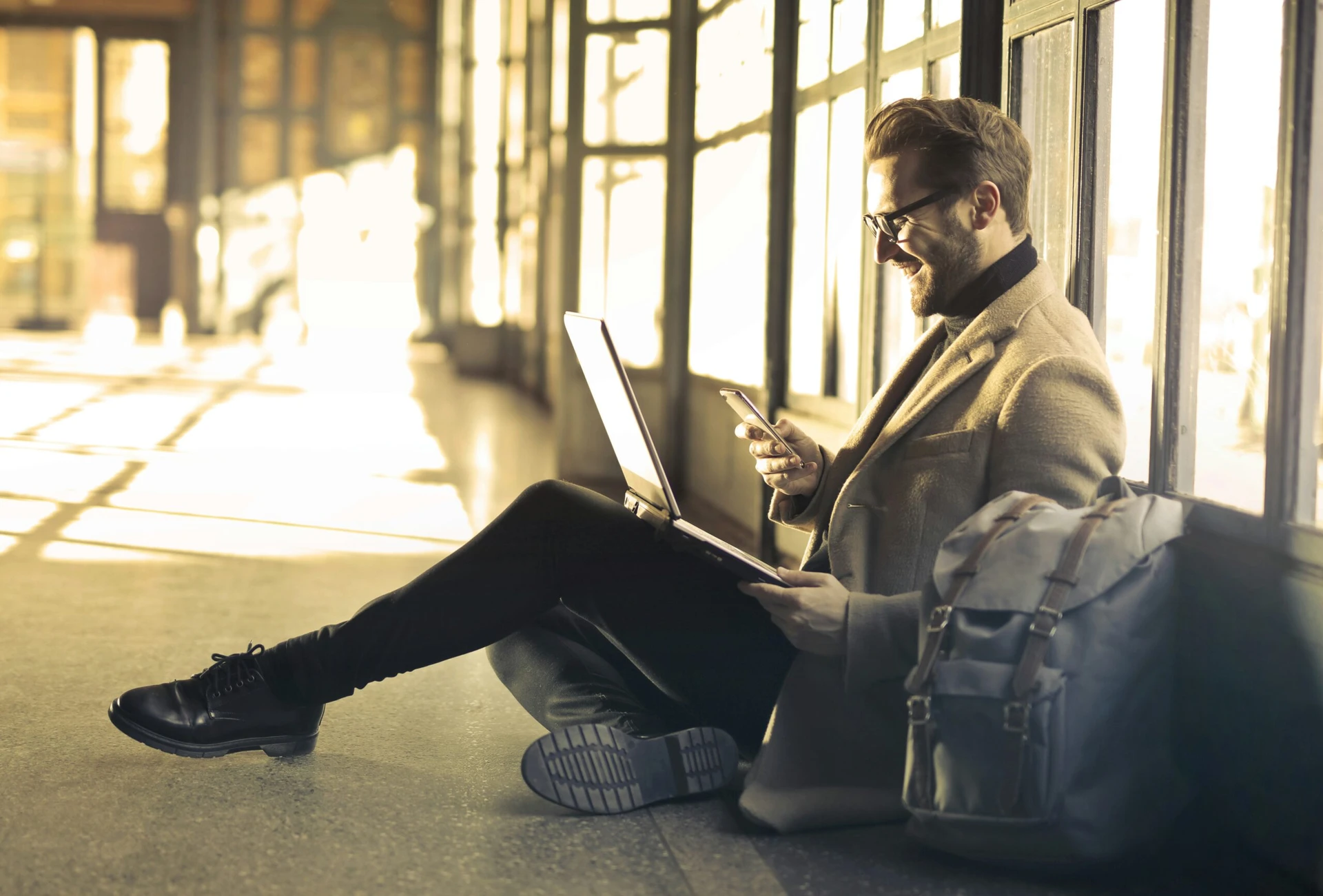 Man sitting on floor by large windows, using a laptop and smartphone, with a backpack beside him.
