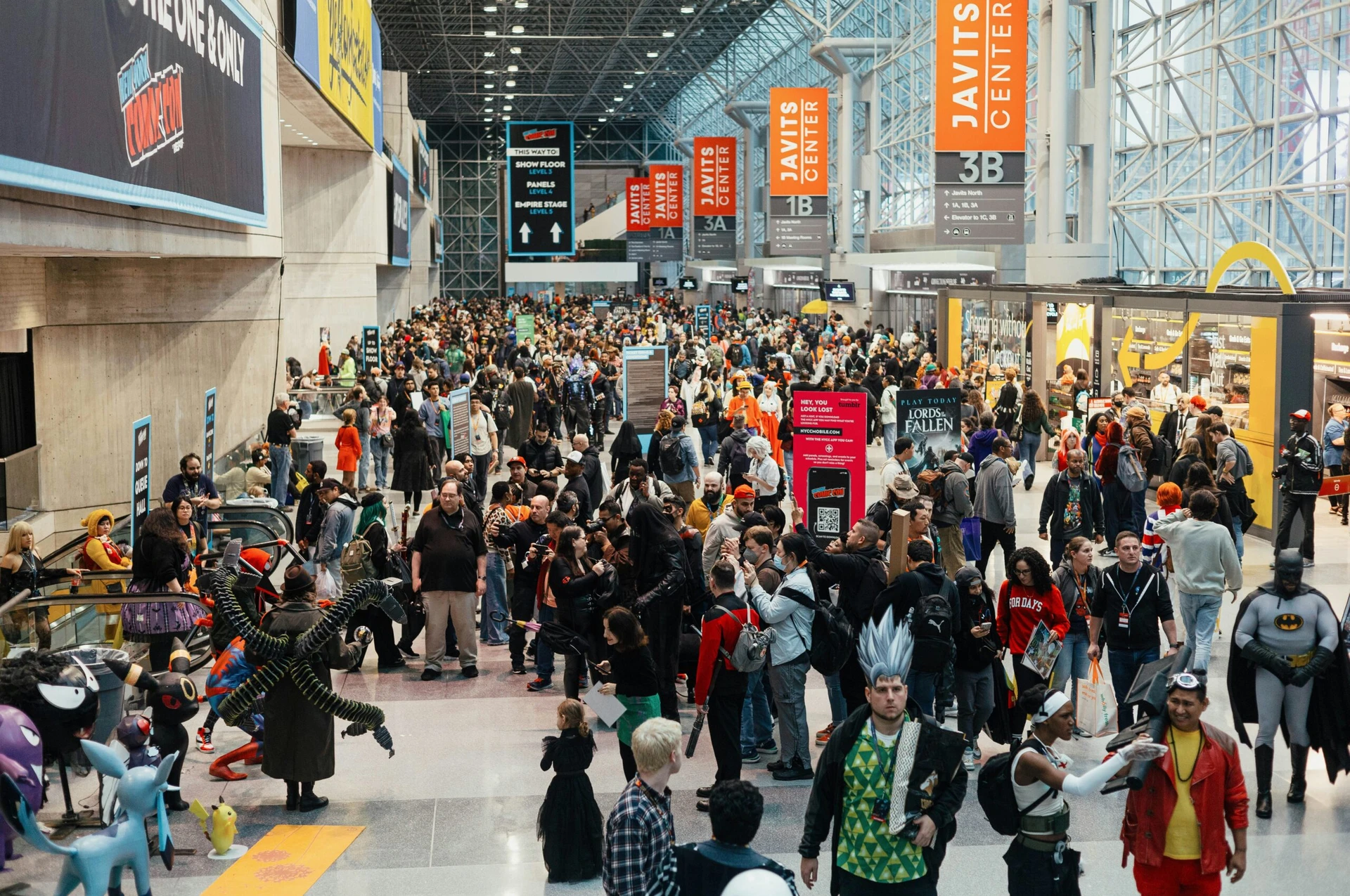 A large crowd of people in costumes gather at a convention center with banners overhead.