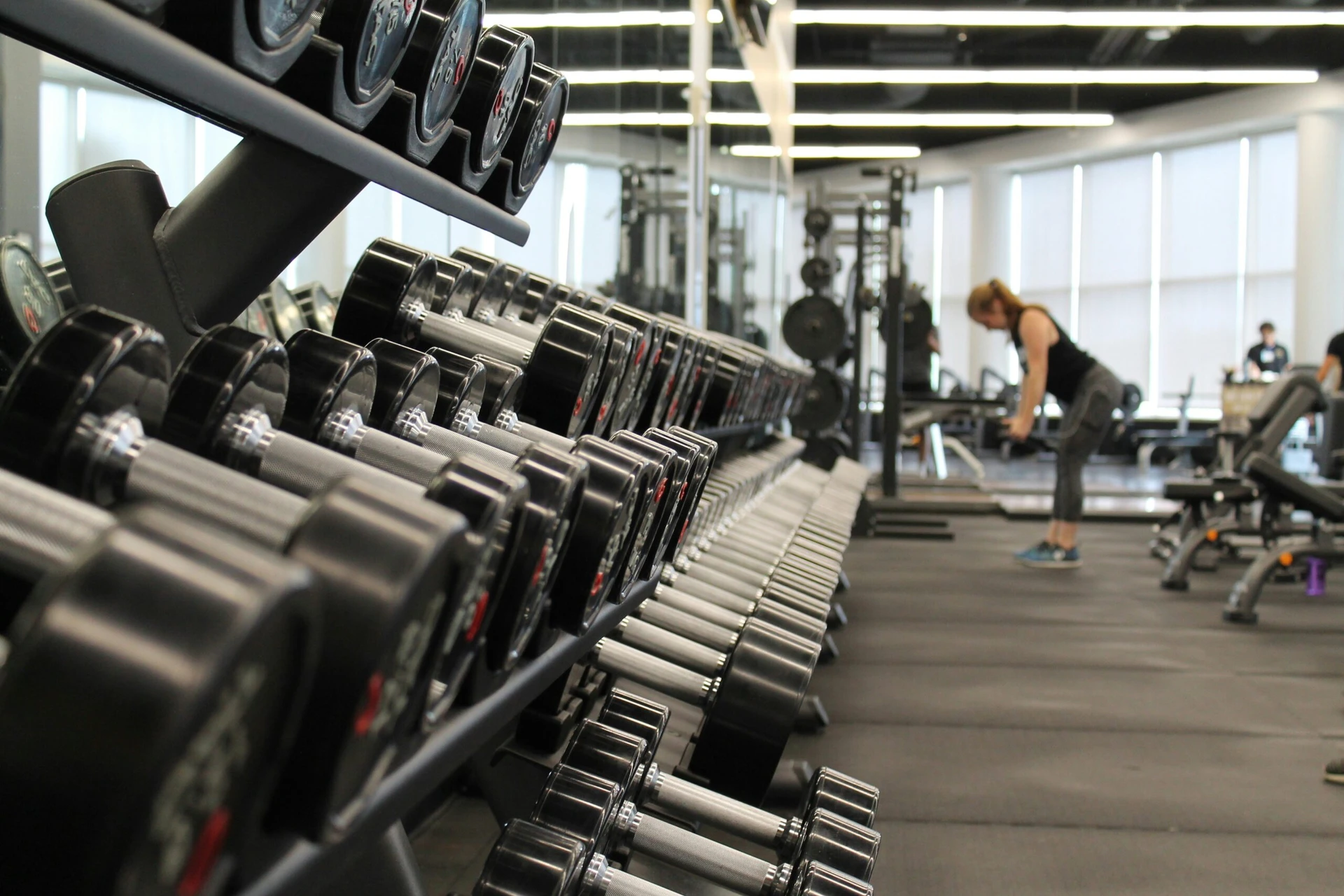 Rows of dumbbells in a gym with people working out in the background.