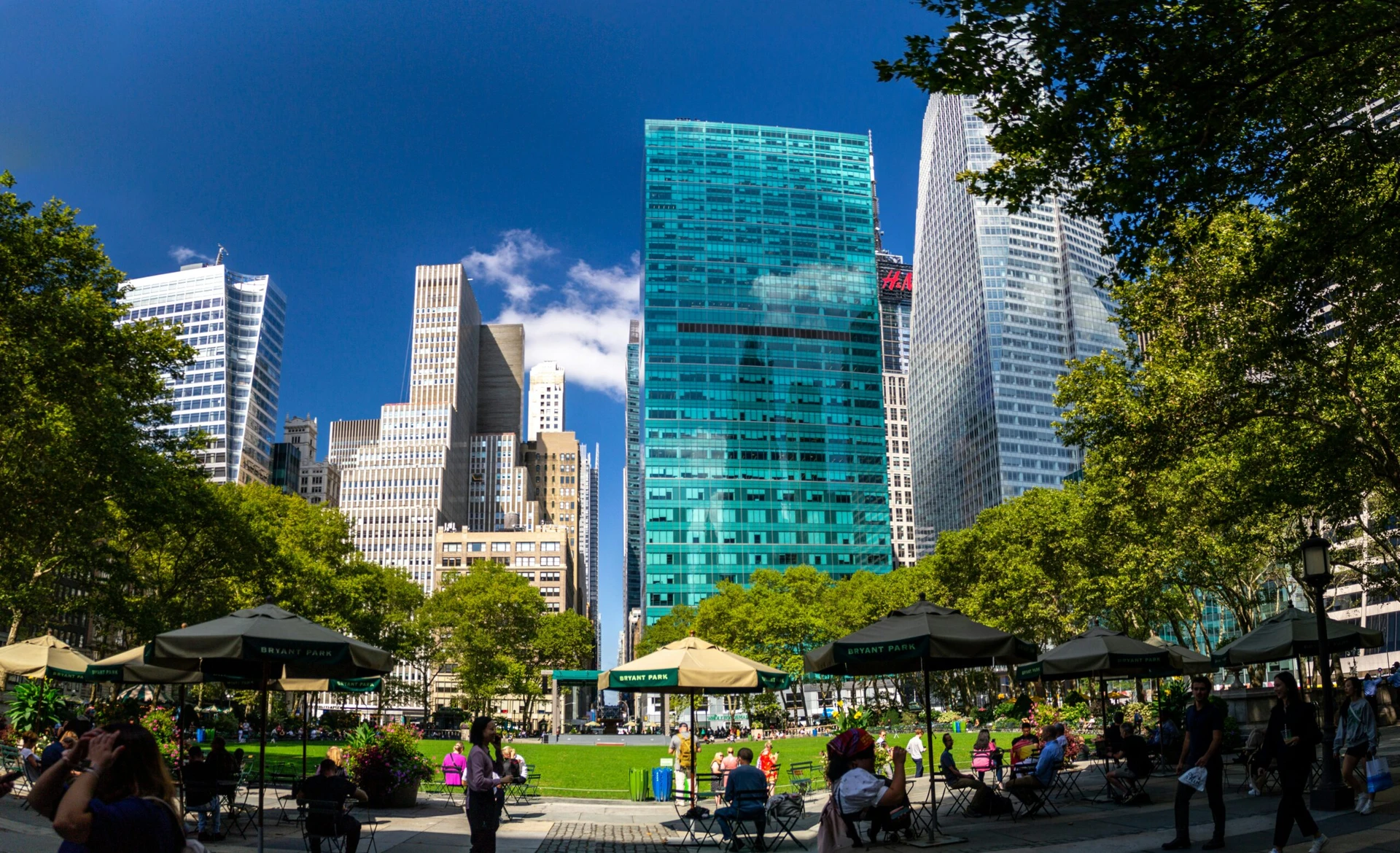 People relax under umbrellas in a city park surrounded by tall skyscrapers on a sunny day.