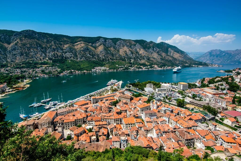 A coastal town with red-roofed buildings, marina, and mountains under a clear blue sky.