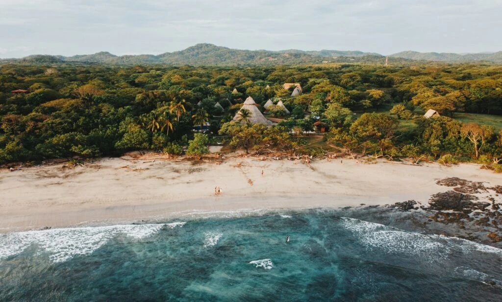 Aerial view of a sandy beach, clear blue water, and lush green jungle with huts in the background.
