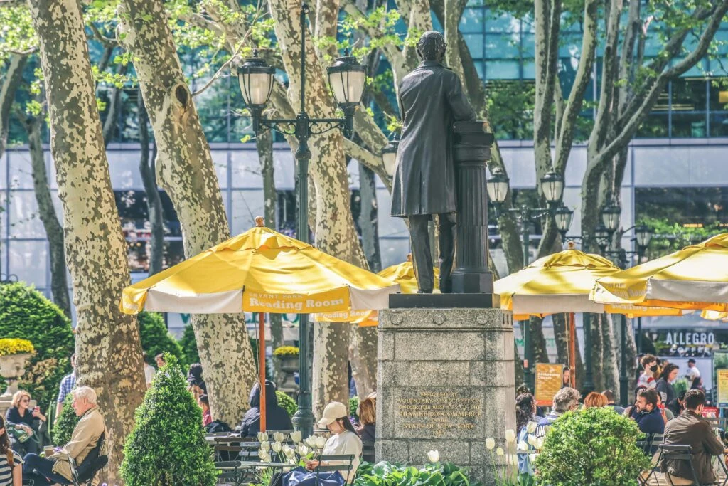 Statue in a busy park with yellow umbrellas, trees, and people sitting at tables on a sunny day.