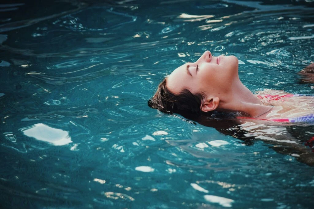 Woman floating on her back in a pool, eyes closed, appearing relaxed in the blue water.