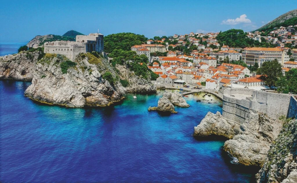 A coastal fortress with red-roofed buildings overlooks clear blue water and rocky cliffs under a sunny sky.