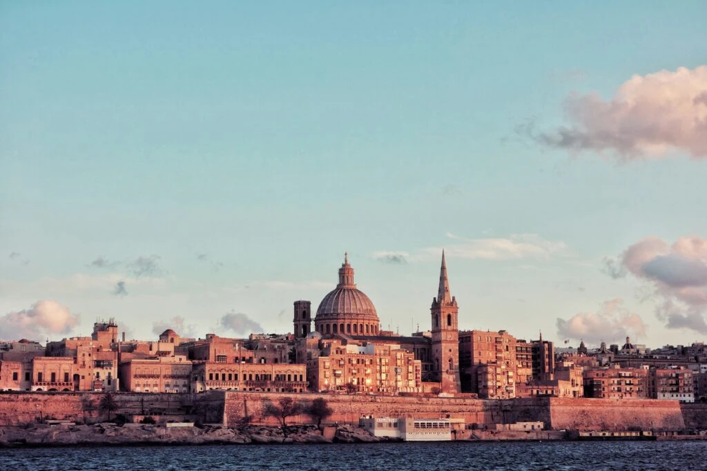 City skyline with historic buildings and a large dome by the water under a partly cloudy sky at sunset.