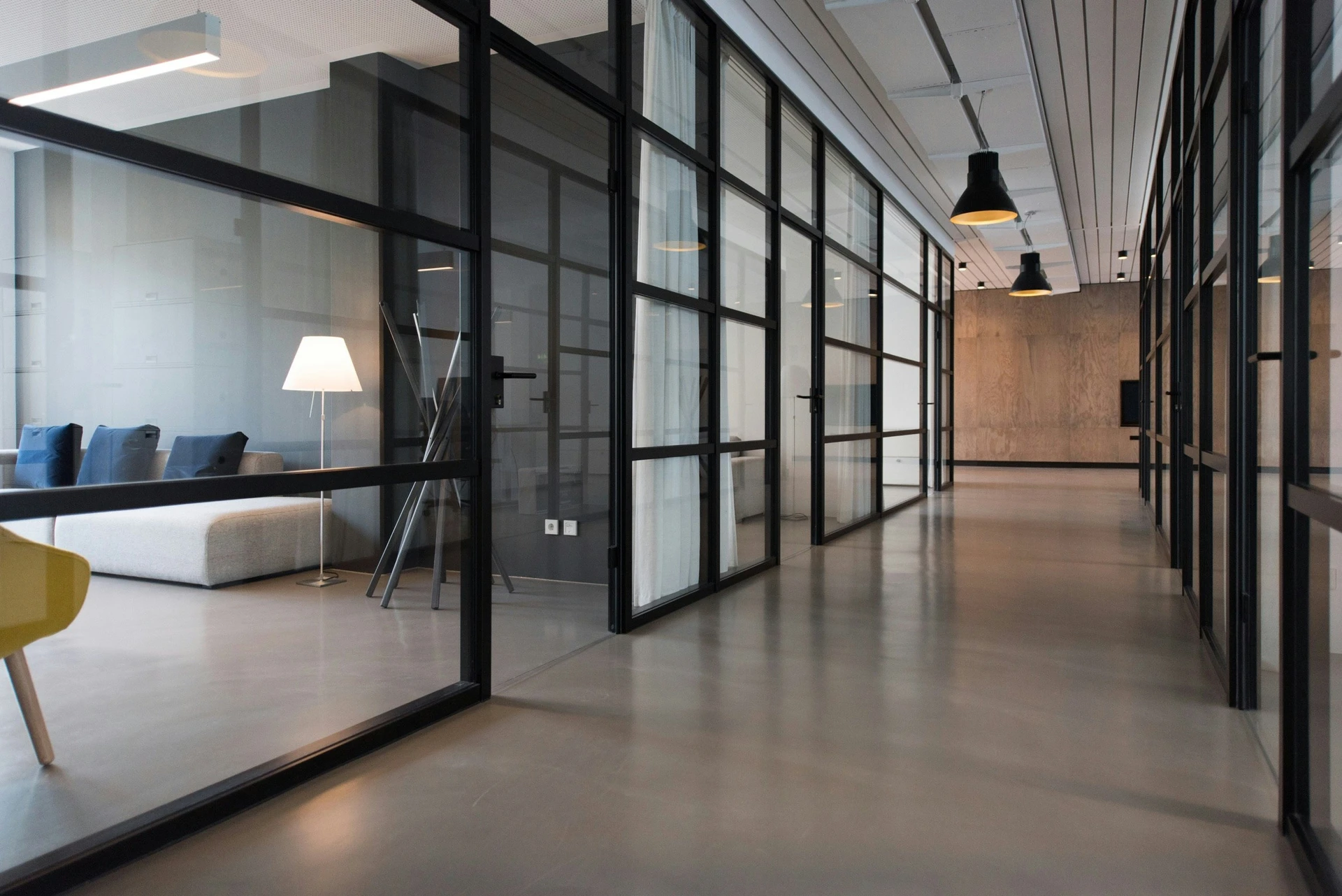 Modern office hallway with glass-walled rooms, a sofa, lamp, and minimalistic decor.