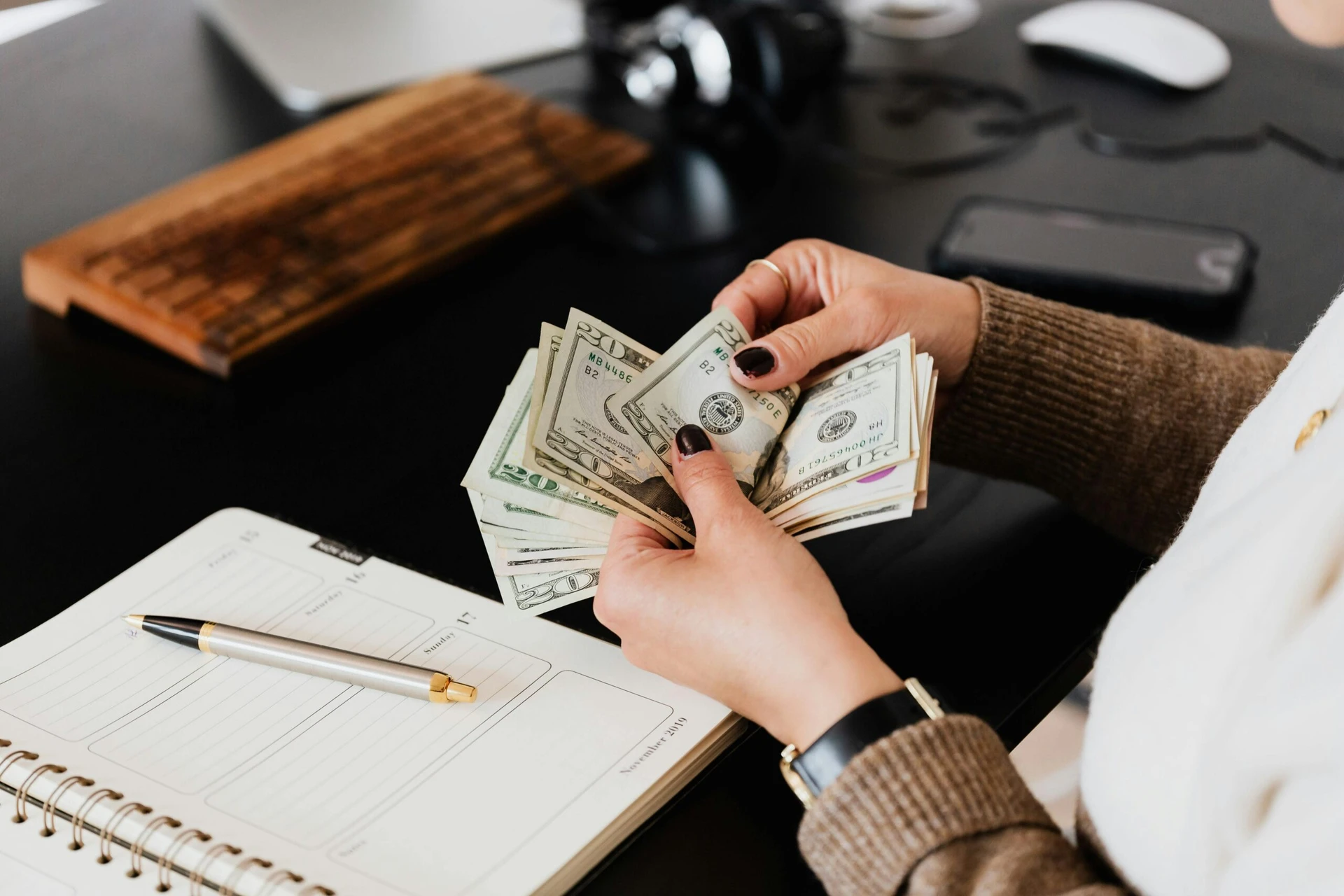 Person counting US dollar bills at a desk with a notebook, pen, keyboard, and phone nearby.