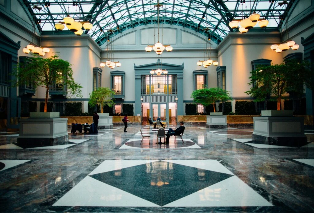 Spacious atrium with marble floors, tall windows, hanging lights, and trees; people sitting and walking.