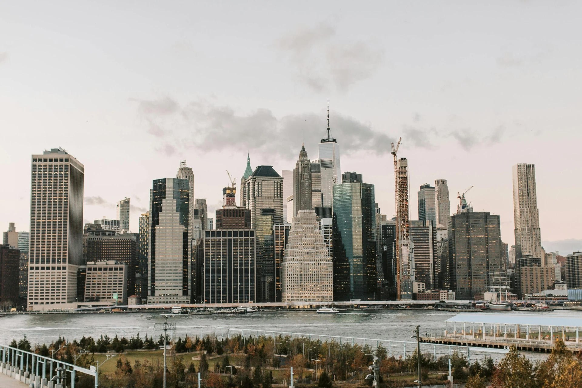 Skyline view of downtown Manhattan buildings with the river and park in the foreground at dusk.