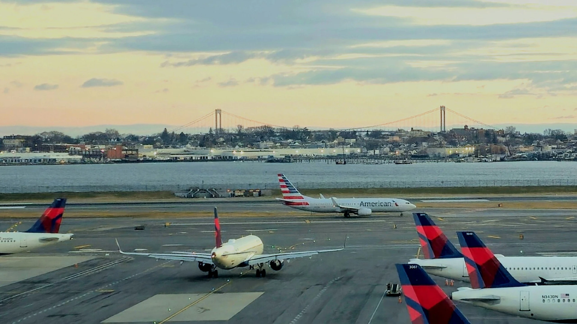Planes on a runway at an airport, with a bridge and water in the background under a cloudy sky.