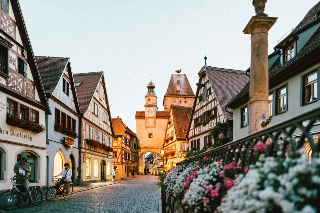 A cobblestone street lined with charming, historic German buildings and colorful flowers at sunset.