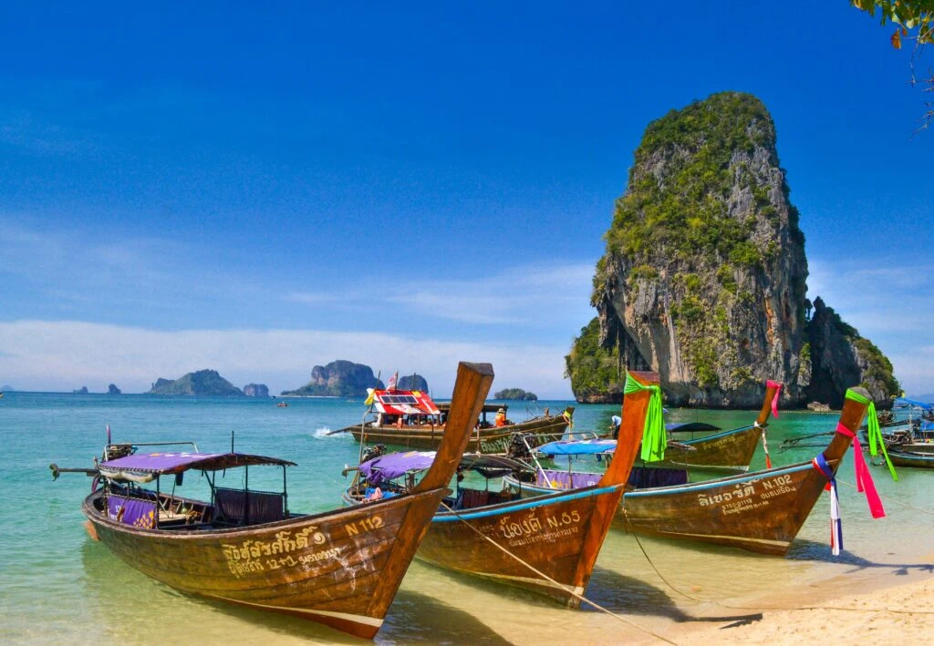 Traditional wooden boats with colorful ribbons docked on a beach with clear water and a limestone cliff.