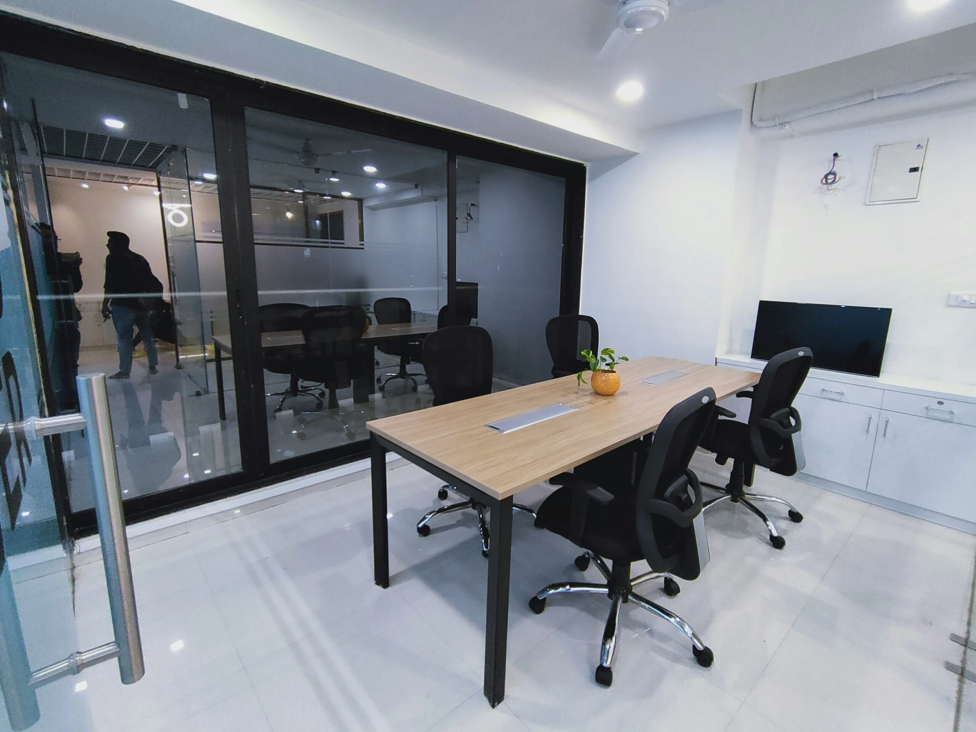 Small modern office with black chairs, wooden desks, a potted plant, and glass walls separating meeting rooms.