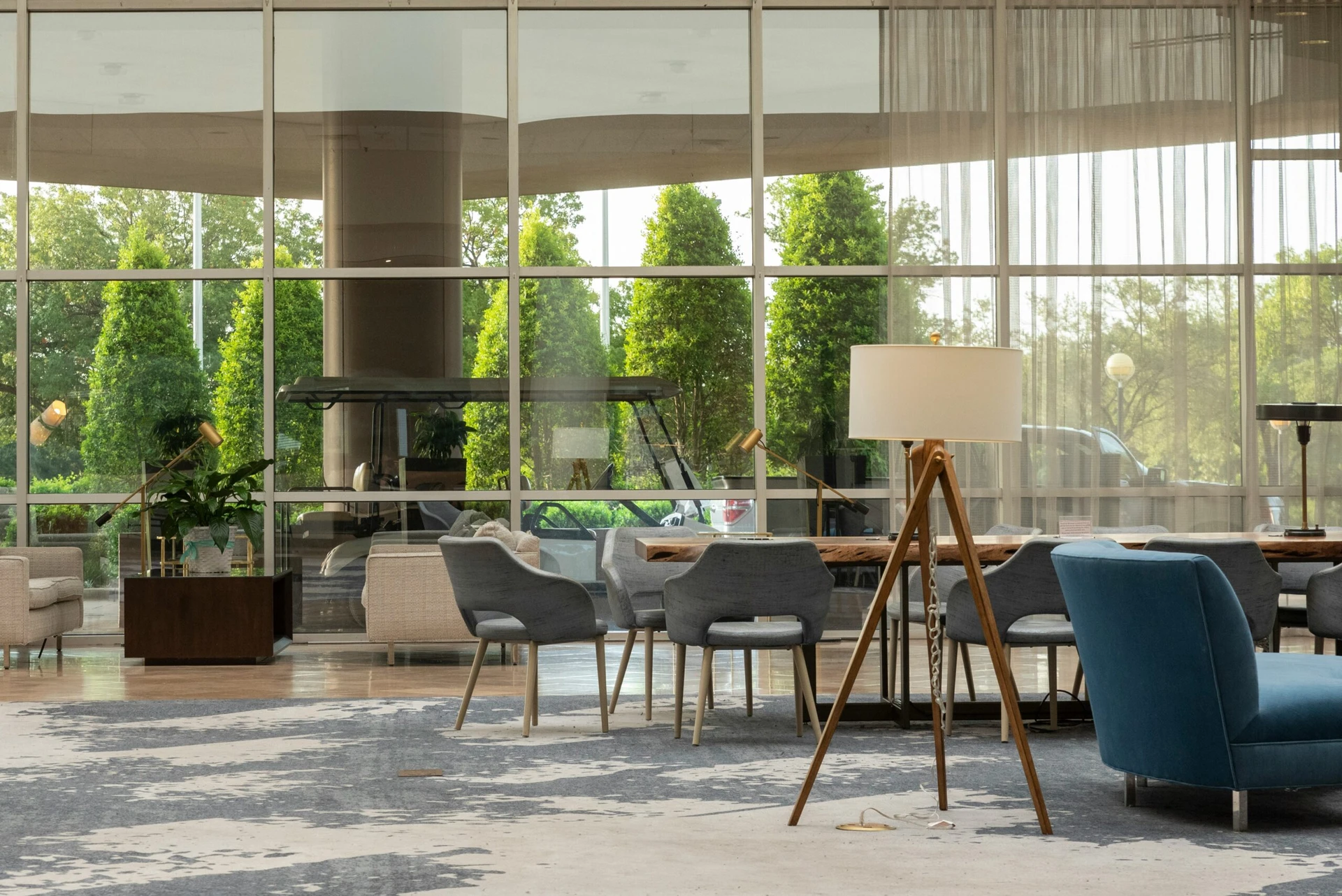Modern hotel lobby with blue and gray chairs, tables, a floor lamp, and large windows showing greenery outside.