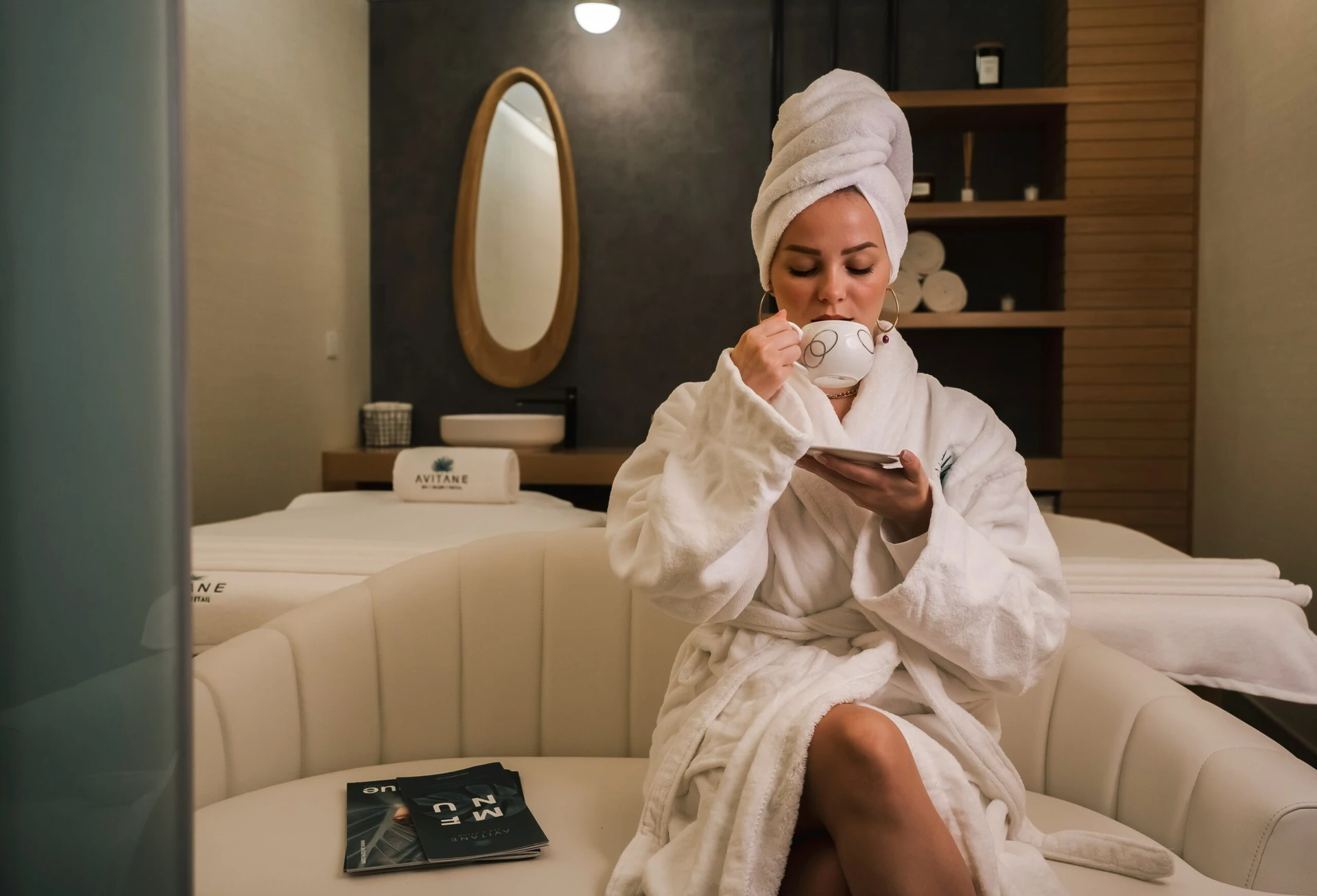 Woman in a white robe and towel enjoys tea in a spa lounge, sitting on a white couch.