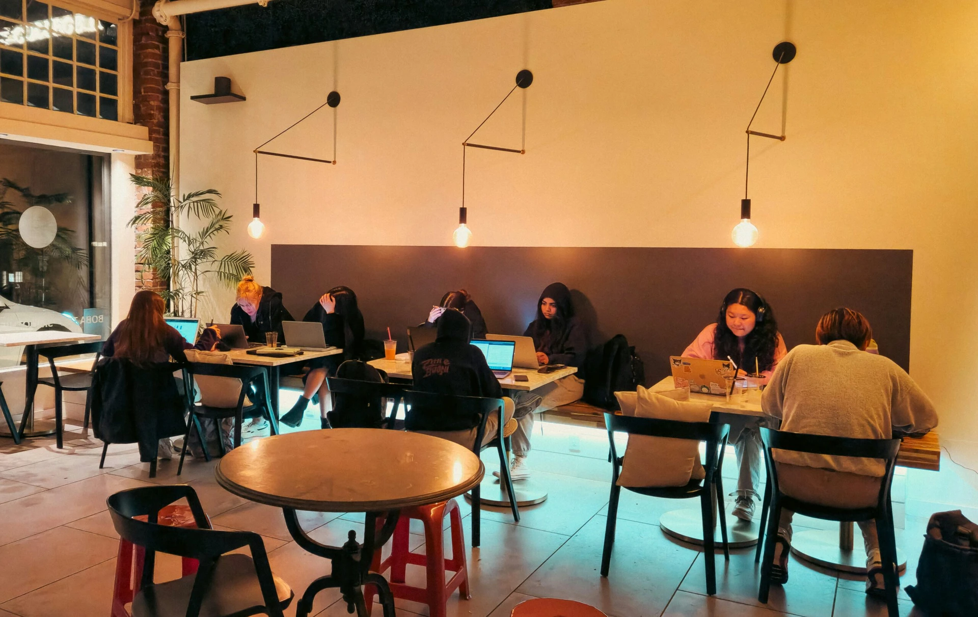 People work on laptops at a well-lit café, sitting at tables against a wall with hanging lights.
