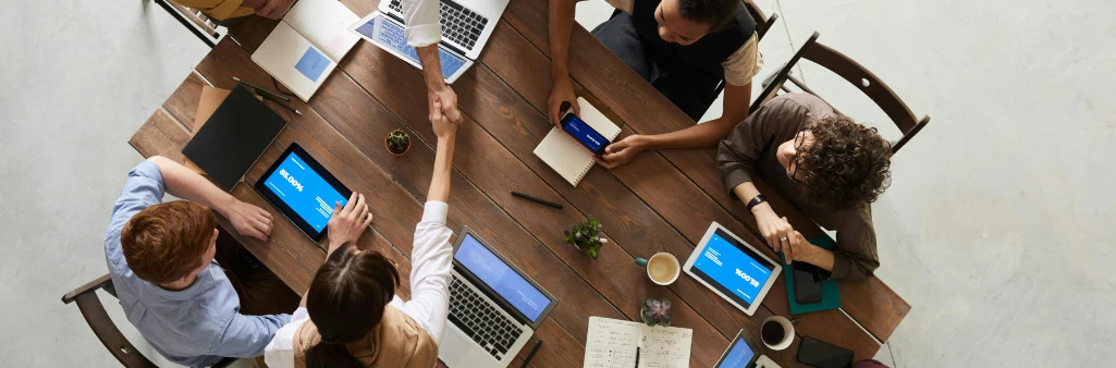 Five people sit around a table with laptops and tablets; two people are shaking hands across the table.