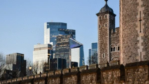 Medieval stone tower and wall in foreground with modern glass skyscrapers in the background under a clear sky.