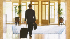 A man in a suit walks indoors, pulling a suitcase and carrying a bag toward glass doors.
