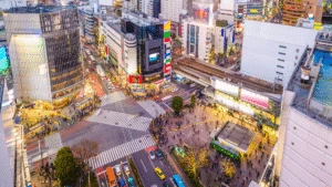 Aerial view of busy Shibuya Crossing in Tokyo with crowds, buildings, and colorful cars at dusk.