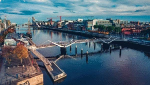 A modern white pedestrian bridge spans a calm river in a vibrant, urban cityscape under a blue sky.