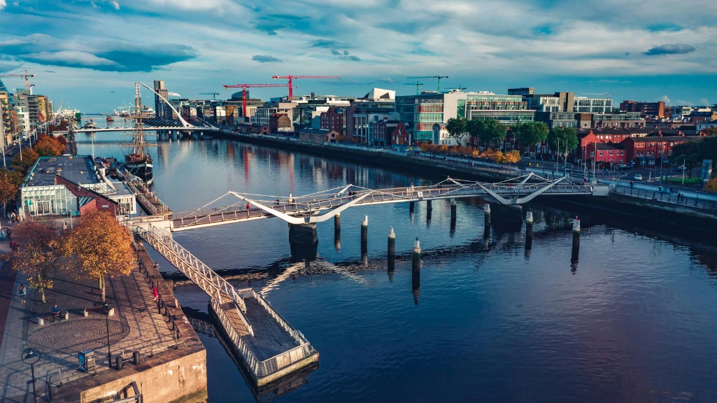 A modern white pedestrian bridge spans a calm river in a vibrant, urban cityscape under a blue sky.