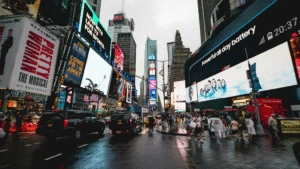 Crowded Times Square on a rainy day, with bright billboards, taxis, and people walking with umbrellas.