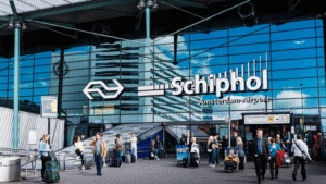 People with luggage outside the glass entrance of Schiphol Amsterdam Airport, with signs and reflections visible.