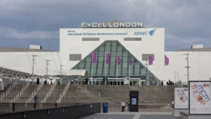 The ExCeL London exhibition centre with glass entrance, steps, and purple flags under a cloudy sky.
