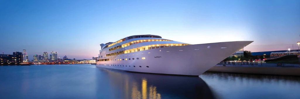 A large, modern white yacht is docked at a city waterfront during dusk with buildings in the background.