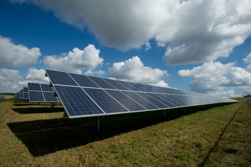 Rows of solar panels on a grassy field under a partly cloudy blue sky.