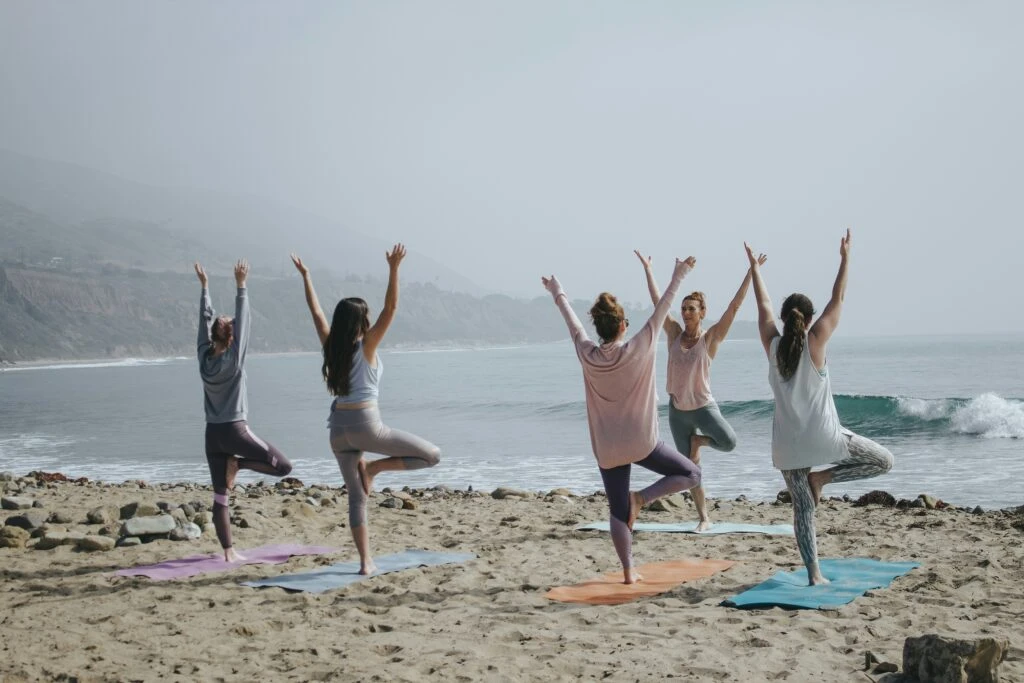 Five people practice yoga in tree pose on mats on a sandy beach by the ocean, with mountains in the background.