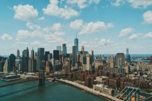 Aerial view of Lower Manhattan skyline with the Brooklyn Bridge and East River under a partly cloudy sky.