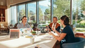 Three people collaborate at a sunlit office table with laptops and paperwork, large windows showing greenery.