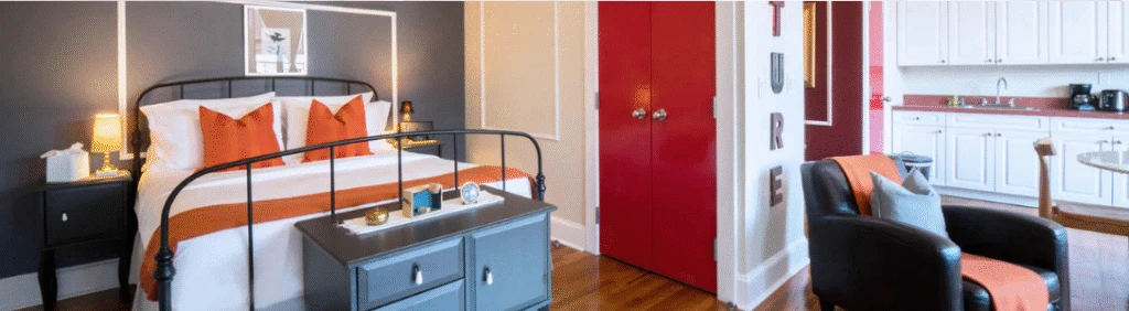 A cozy bedroom with orange accents, a black metal bed, and an adjoining white kitchen area.